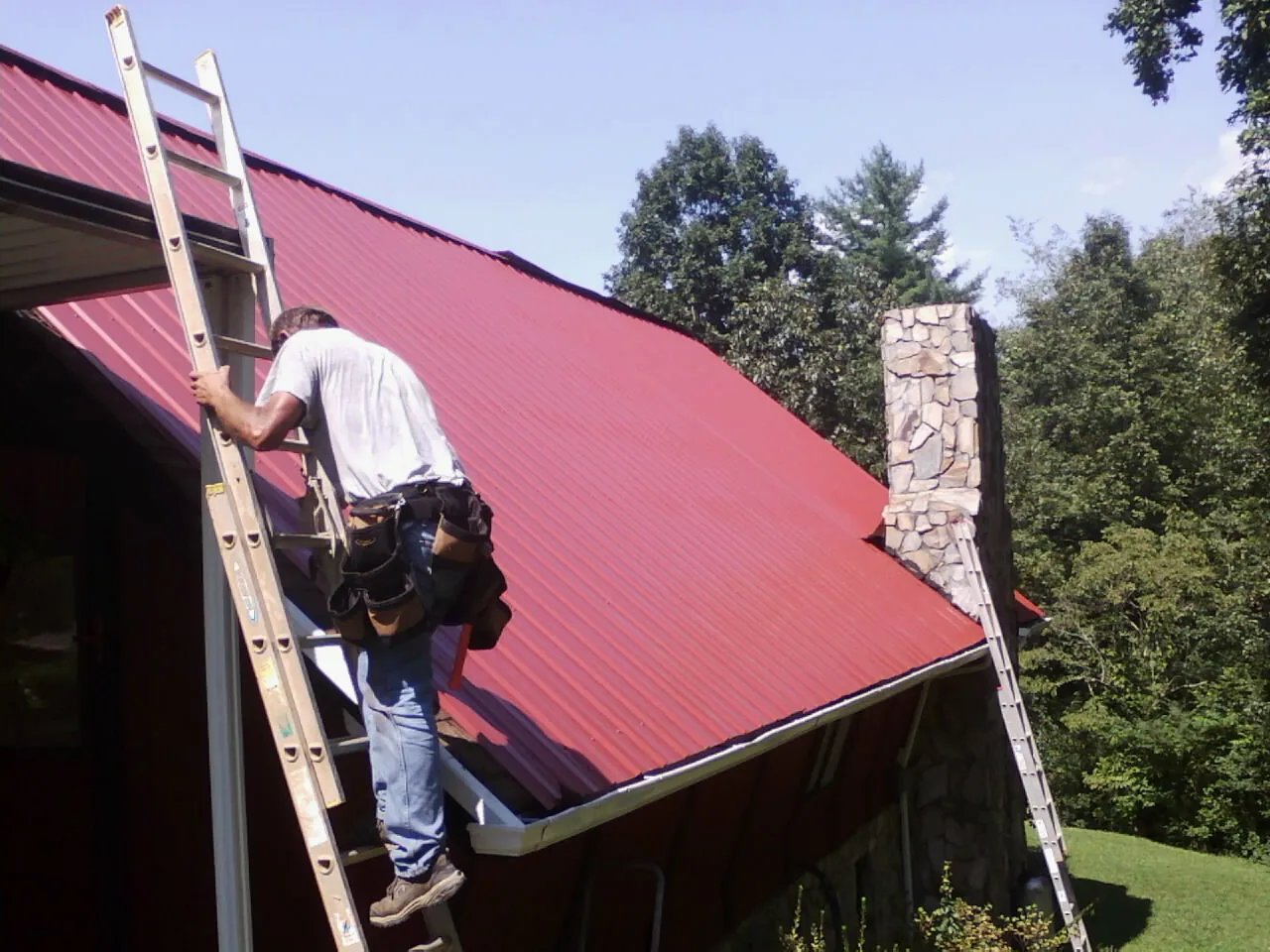 Roofer on a ladder beside a red metal roof and stone chimney — Soffit & Fascia Repair by Roofing Brothers in Herkimer